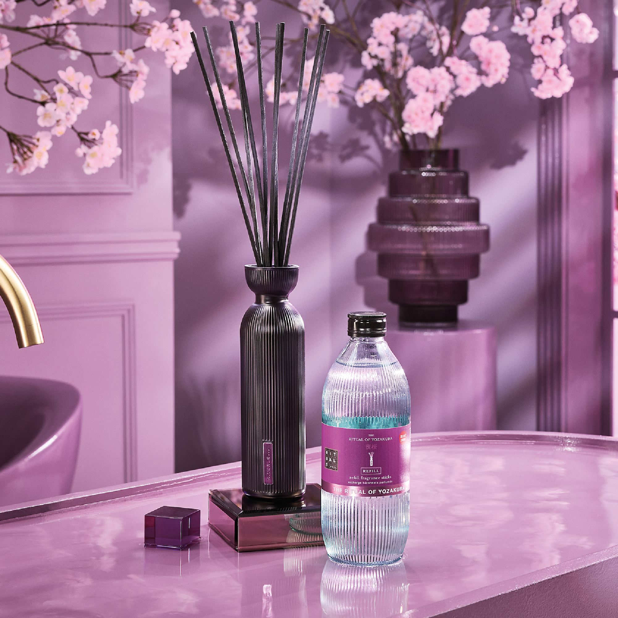 Duftpinner i en svart flaske fra Rituals står på et rosa baderomsbord sammen med en refillflaske merket “THE RITUAL OF YOZAKURA”, “REFILL” og “refill fragrance sticks”. Rosa kirsebærblomster vises i bakgrunnen.