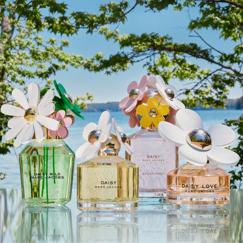 Fyra parfymflaskor från Marc Jacobs kollektion "Daisy" placerade på ett bord utomhus med en sjö och grönskande träd i bakgrunden. Flaskorna har blomformade lock i olika färger och är märkta med "Daisy Wild", "Daisy", "Daisy Eau So Fresh" och "Daisy Love".