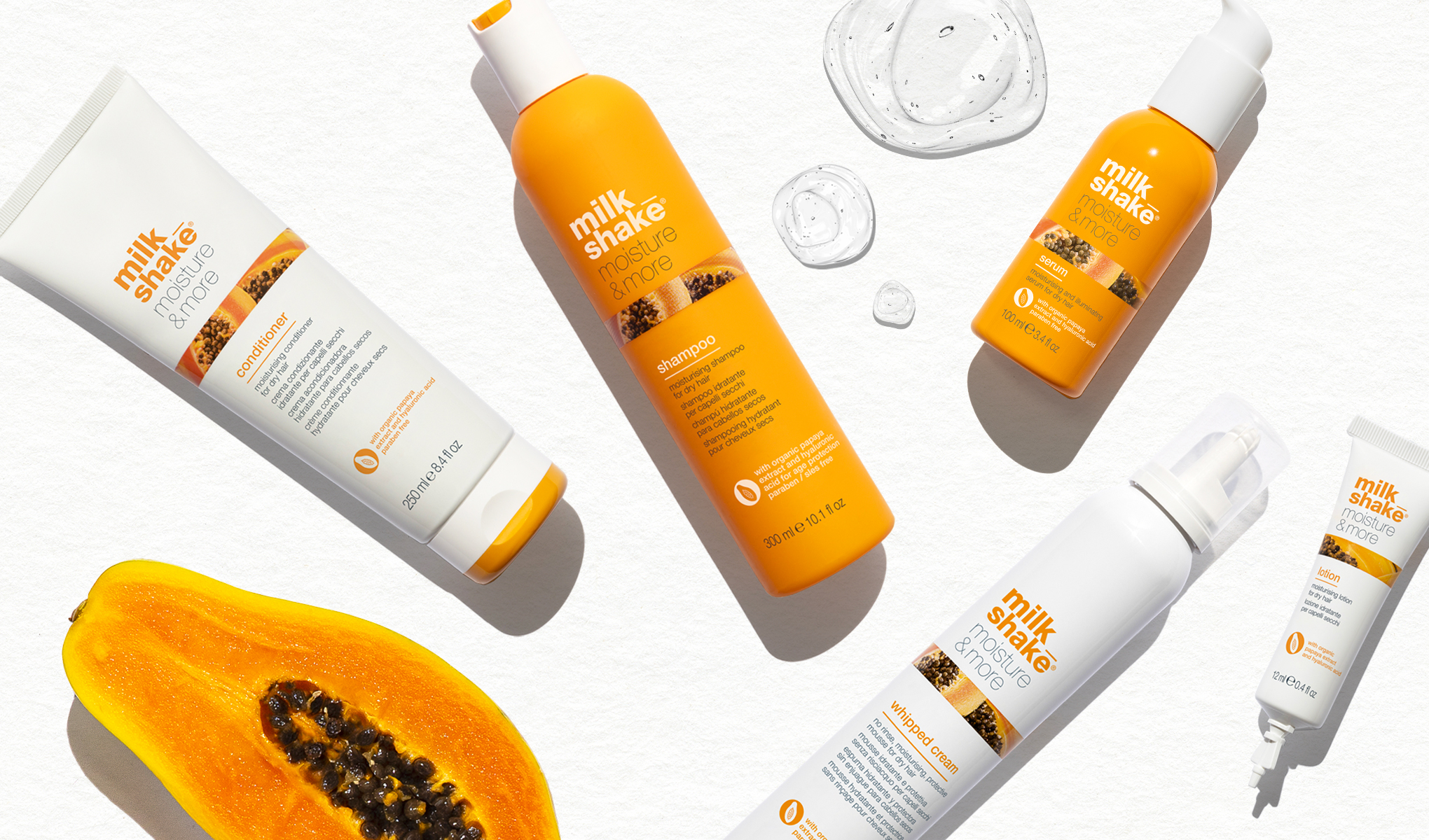 Flera Milk Shake-produkter, såsom schampo, balsam, serum, skum och kräm, placerade på en vit bakgrund. Bilden inkluderar även en halv papaya.