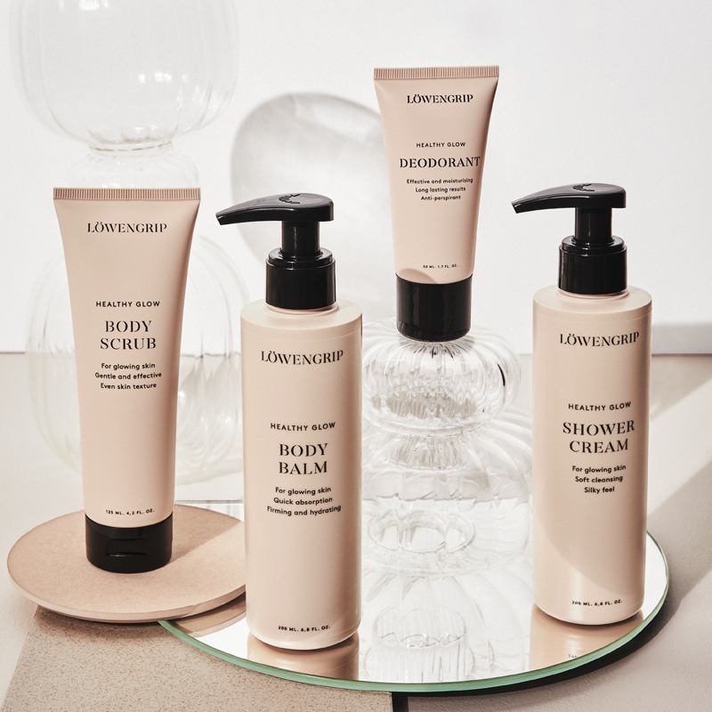 Fire kroppspleieprodukter fra Löwengrips Healthy Glow-serie er stilt opp mot en lys bakgrunn med dekorative glasskupler. Alle produktene har beige emballasje med svart tekst og svarte lokk. Produktene er plassert på et rundt speil og en beige plate.