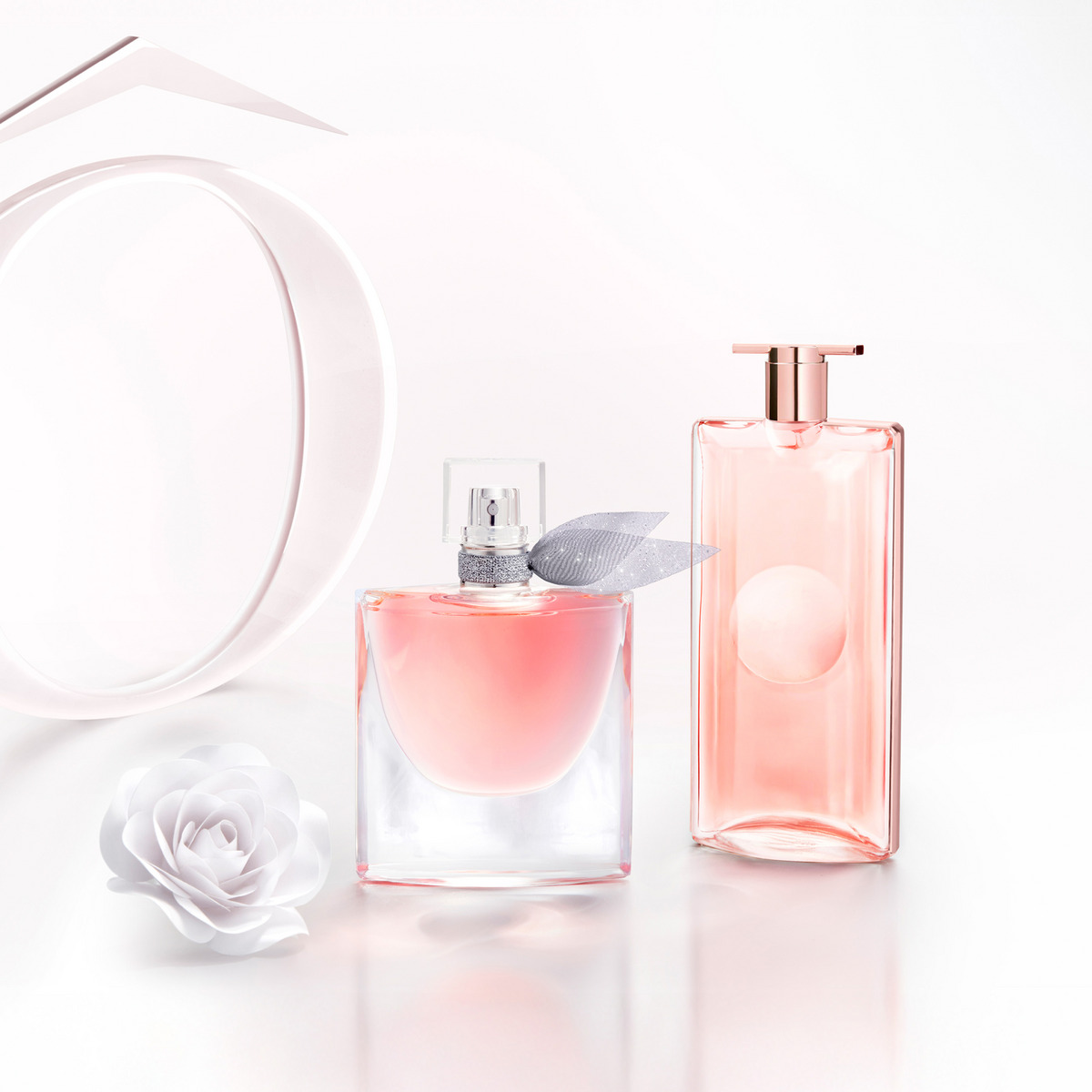 Två rosa parfymflaskor mot ljus bakgrund, den ena med silverfärgad rosett runt halsen, och en vit blomma bredvid.