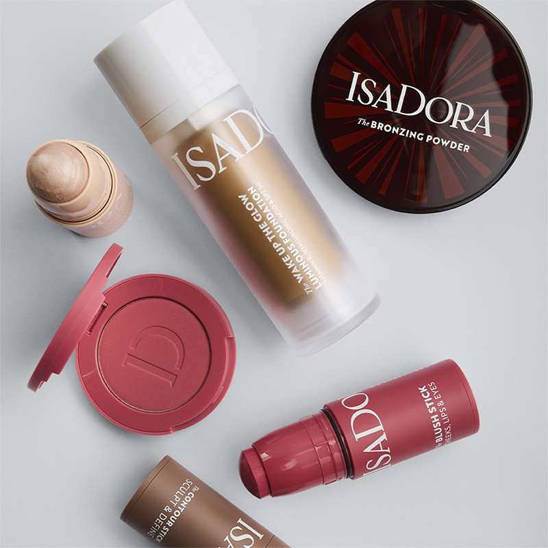 Flere IsaDora makeup-produkter inkludert The Bronzing Powder, The Wake Up the Glow Luminous Foundation og The Blush Stick arrangert mot lys bakgrunn. Synlige tekster på emballasjen viser produktnavn og merke.