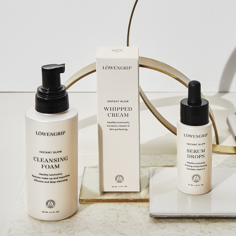 Tre hudpleieprodukter fra Löwengrips Instant Glow-serie er stilt opp mot en lys bakgrunn. Alle produktene har beige emballasje med svart tekst og svarte lokk. Produktene er plassert på ulike tallerkener, og en dekorativ gullring er synlig i bakgrunnen.