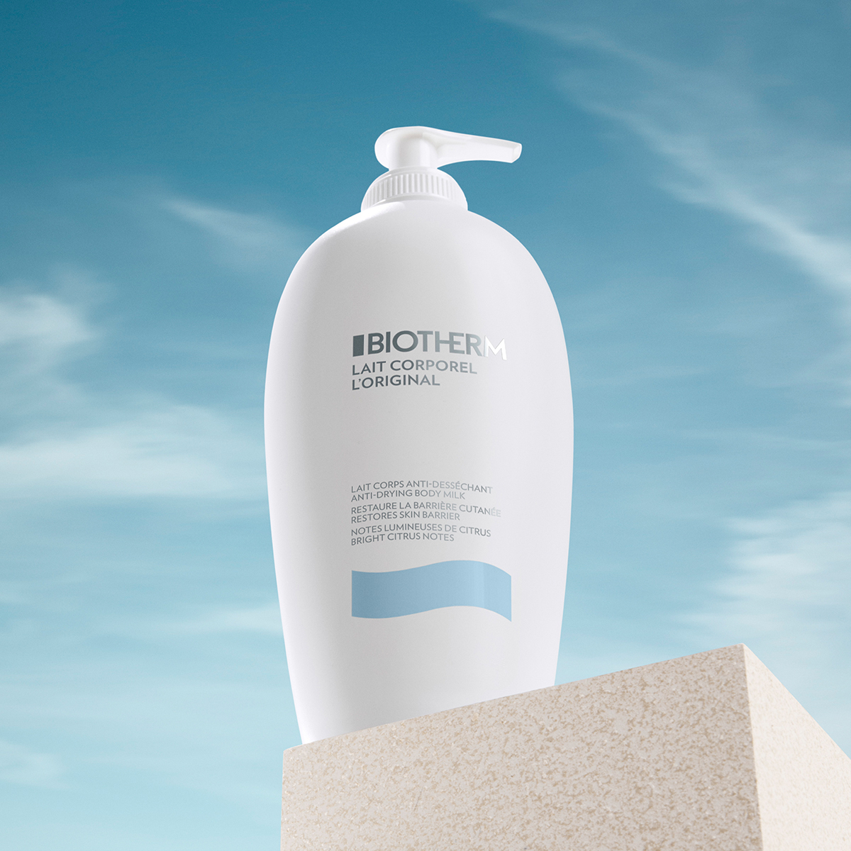 Biotherms Lait Corporel L'Original. Kroppsmjölken kommer i en vit pumpflaska och har texten "Biotherm Lait Corporel L'original" på framsidan. Flaskan står på en beige plattform mot en blå himmel med några vita moln.