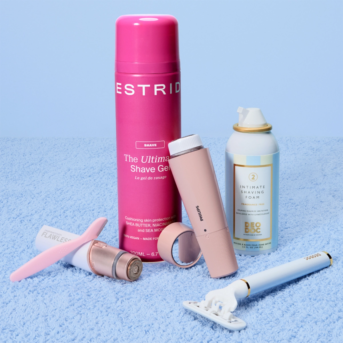 Barberings- og intimpleieprodukter er stilt opp mot en lys blå bakgrunn, blant annet Estrid The Ultimate Shave Gel, DeoDoc Intimate Shaving Foam Fragrance Free, Philips elektrisk trimmer og Flawless hårfjerner. Produktene vises i rosa, hvite og pastellfargede emballasjer sammen med barberhøvel og groomingverktøy for intimbarbering og hudpleie.
