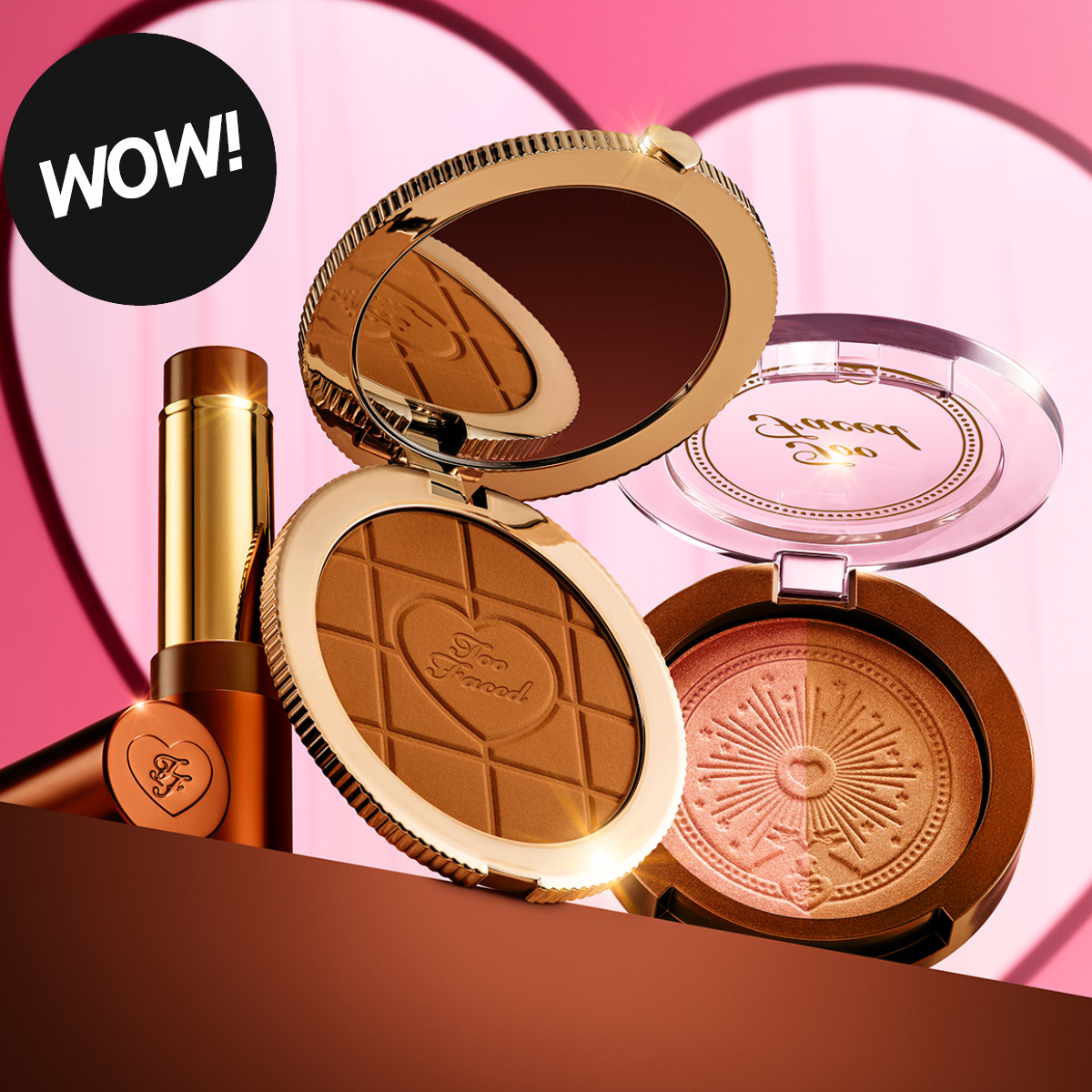 Too Faced -meikkituotteita vaaleanpunaisilla tasoilla sydänaiheisilla yksityiskohdilla: avoin aurinkopuuteri, avoin hohdepuuteri ja kultainen huulipuna. Tuotteissa näkyy Too Faced -logo ja vasemmassa yläkulmassa musta merkki tekstillä "WOW!".