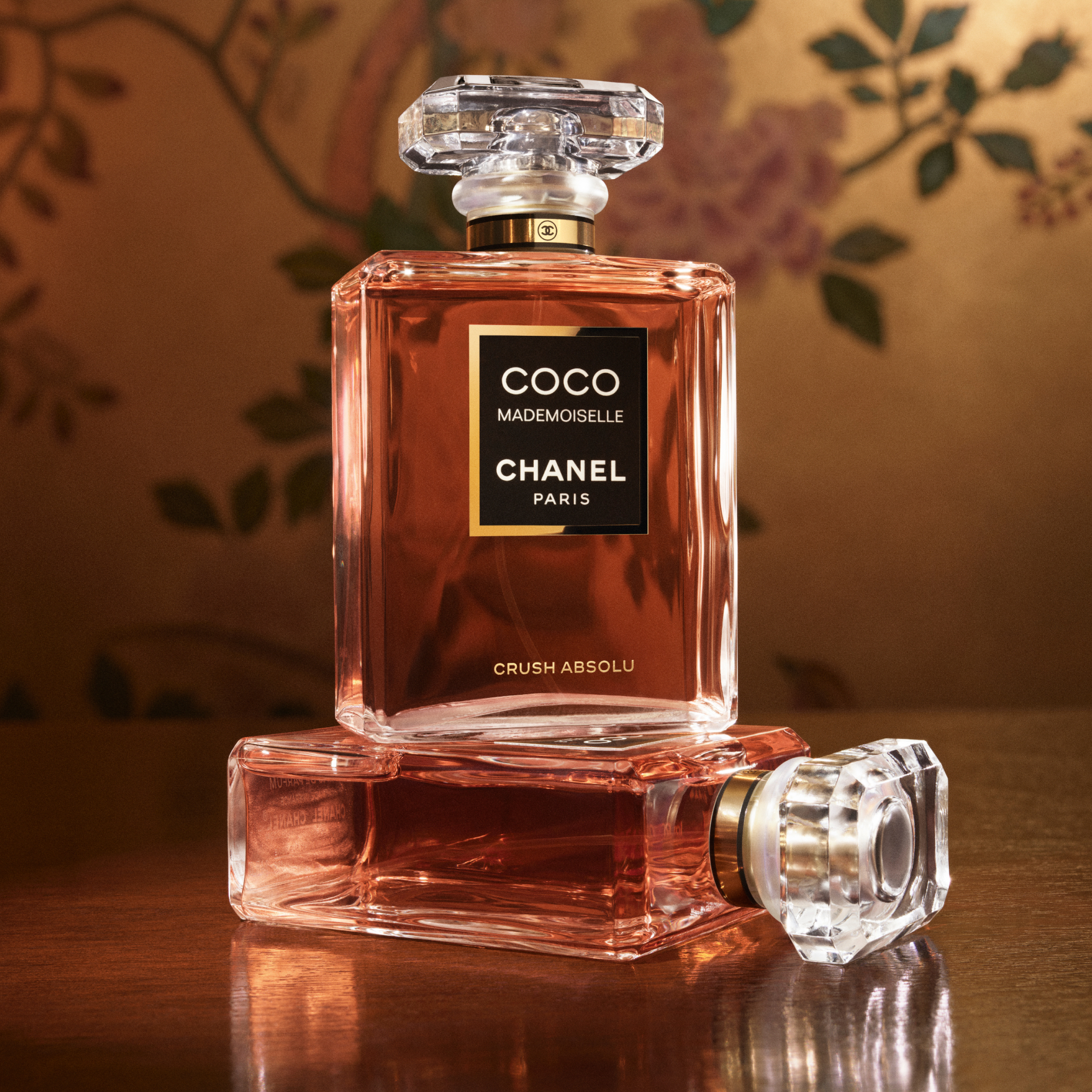 To CHANEL Coco Mademoiselle Crush Absolu-parfymeflasker mot en varm, blomstrete bakgrunn.