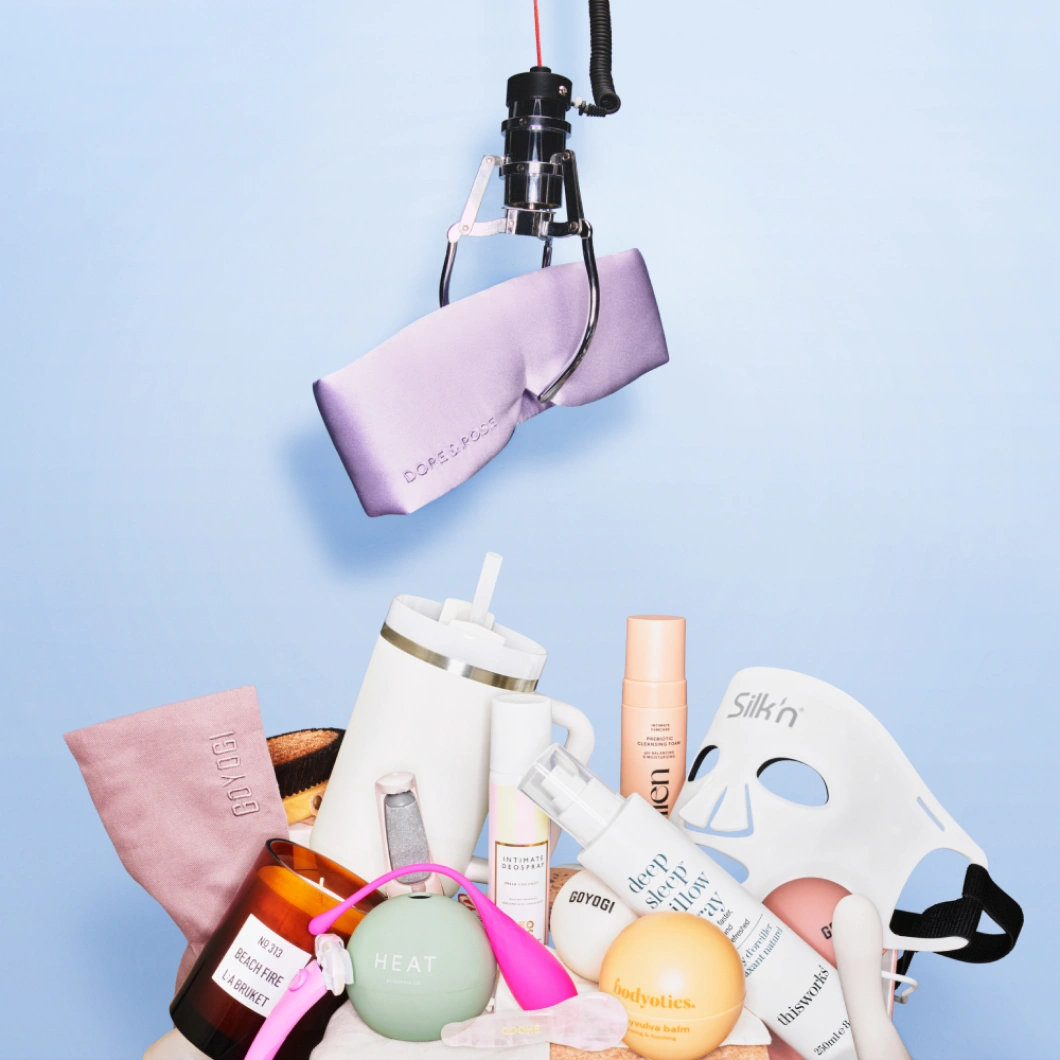 En kloarm håller ett lila sidenliknande tyg ovanför olika wellnessprodukter mot ljusblå bakgrund. Produkterna inkluderar Silkn LED-mask, dryckesmugg, kroppsvård och wellnessaccessoarer.