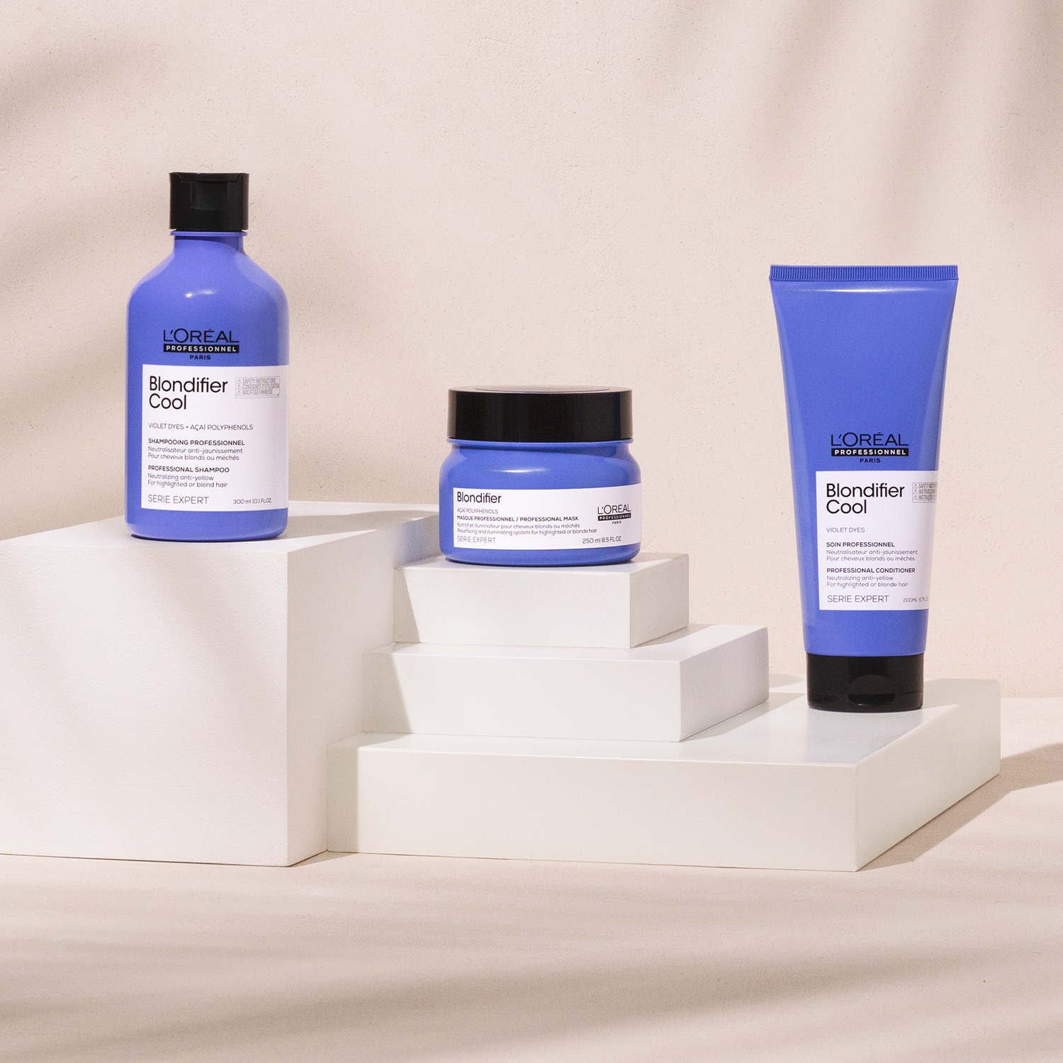 Tre hårpleieprodukter fra L'Oréal Professionnel Blondifier Cool-serien er stilt opp på hvite kuber mot en beige bakgrunn. Alle produktene har blå emballasje med hvite etiketter og svarte lokk.