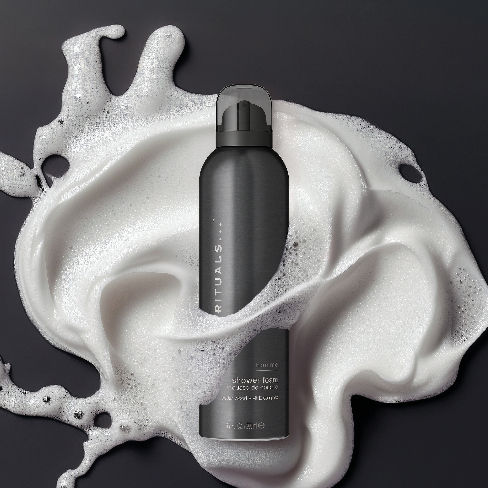 Svart flaske med Rituals Homme shower foam står i hvitt dusjskum mot mørk bakgrunn. På flasken står tekstene “RITUALS”, “homme”, “shower foam” og “cedar wood + vit E complex”.