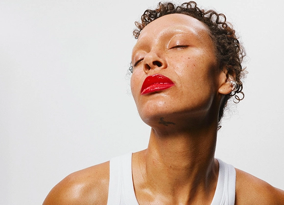 Puoliprofiilinen muotokuva henkilöstä vaaleaa taustaa vasten. Henkilöllä on lyhyet kiharat hiukset, suljetut silmät ja punainen huulipuna. Hartiat ja ylävartalon osa näkyvät, ja henkilöllä on valkoinen hihaton paita.