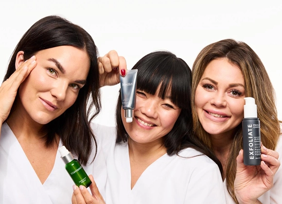 Tre smilende kvinner i hvite frakker holder opp hudpleieprodukter fra Exuviance, Medik8 og Paula’s Choice mot en hvit bakgrunn.