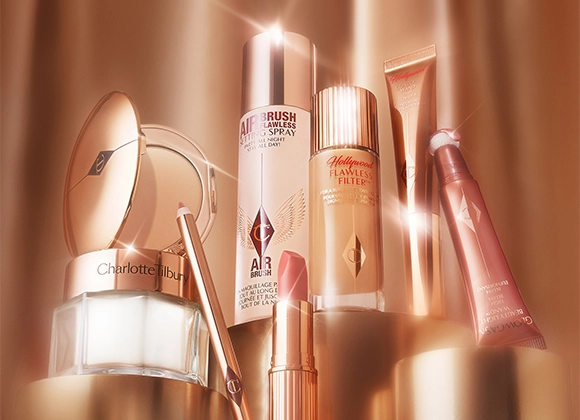 En samling av Charlotte Tilbury skjønnhetsprodukter, inkludert Airbrush Flawless Setting Spray, Hollywood Flawless Filter, Hollywood Contour Wand, Glowgasm Beauty Light Wand, en leppeblyant og en pudderkompakt. Produktene er elegant plassert og belyst med et varmt lys.