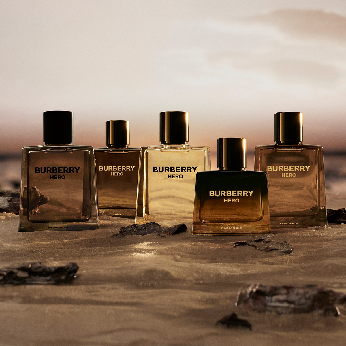 Fem Burberry Hero-duftflasker plassert på sand ved vannkanten i solnedgang.