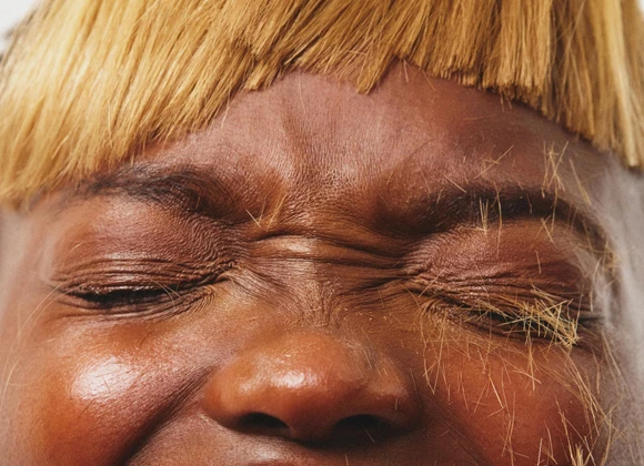 Nærbilde av et ansikt med lukkede øyne og rynket panne. Personen har en lys blond, rettklippet lugg og avklippede hårstrå liggende på øyelokkene.