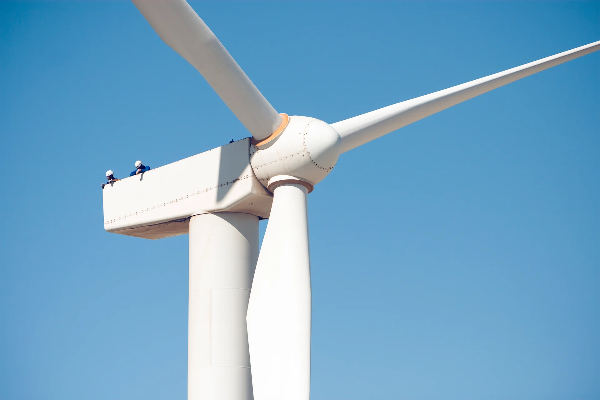 This image shows two workers atop a large wind turbine, performing maintenance or inspection. The turbine’s blades are prominently visible against a clear blue sky, highlighting the scale of wind energy infrastructure and the importance of safety and technical expertise in the wind industry.