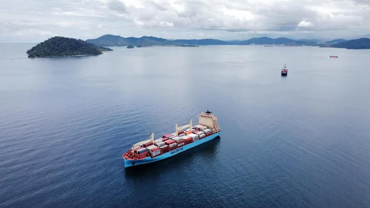 A ship at sea carrying containers, with mountains and other vessels visible in the background, illustrating global trade and maritime logistics amidst a scenic landscape.