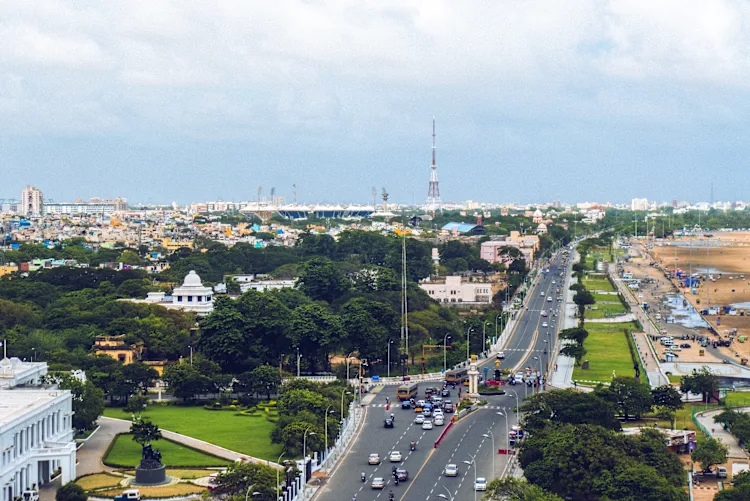An aerial view of Chennai, featuring a bustling highway alongside lush green spaces and the Marina Beach shoreline, capturing the city's vibrant blend of urban life and coastal charm.