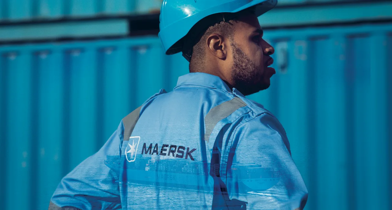 A Maersk worker in a blue uniform and hard hat stands in front of containers, symbolising professionalism and dedication in the shipping and logistics industry.