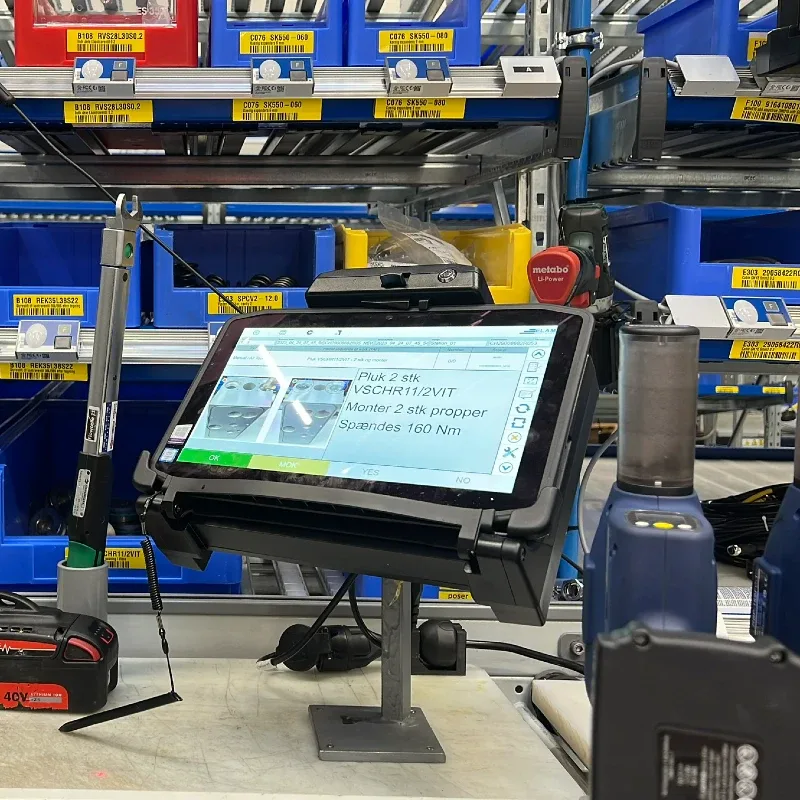 Tablet på et stativ viser instruktioner i et lager med blå kasser og værktøj i baggrunden.