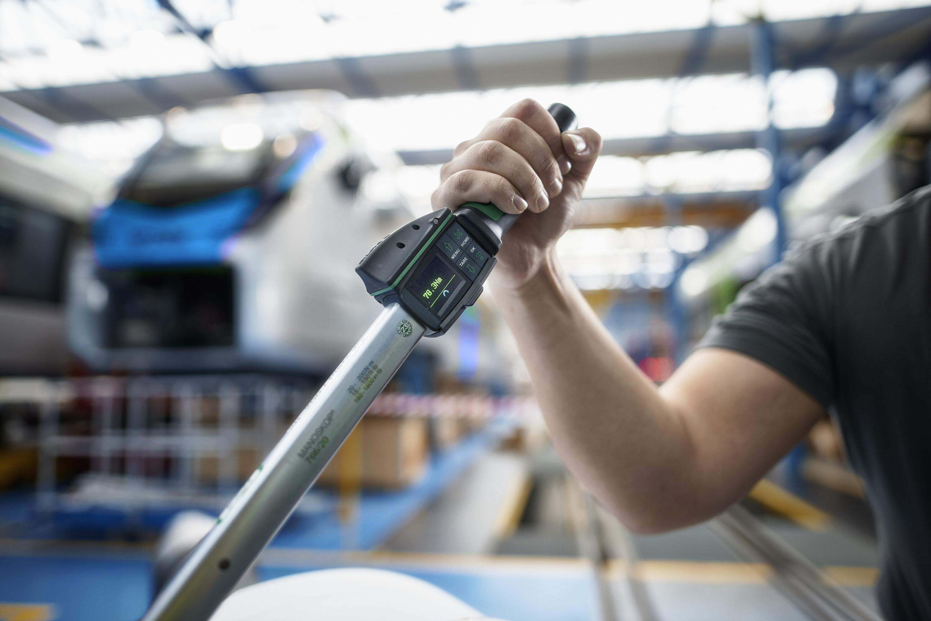 Persona usando una llave dinamométrica digital. El par es visible en una pequeña pantalla. El fondo desenfocado muestra un tren en una fábrica de construcción ferroviaria.