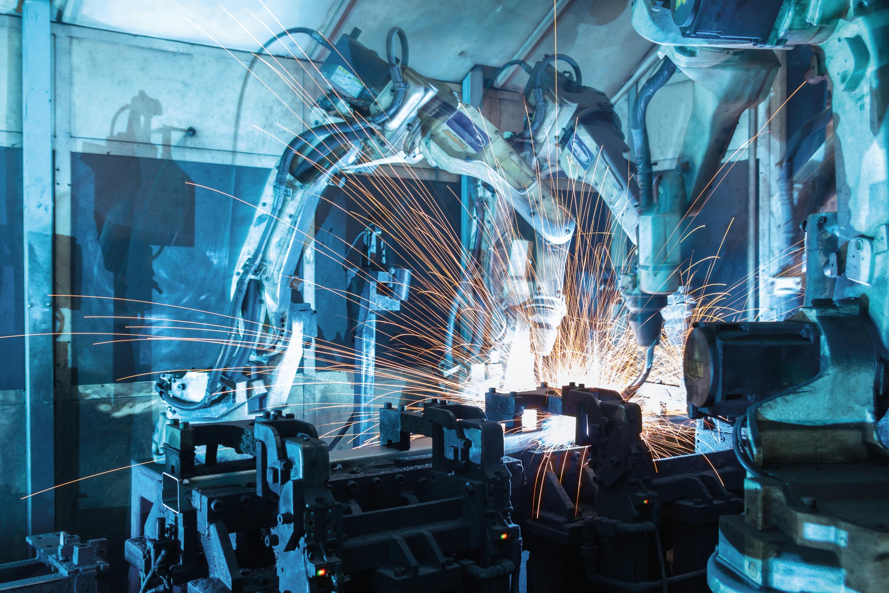 Industrial robotic arms welding metal with bright orange sparks flying in a blue-lit factory setting.