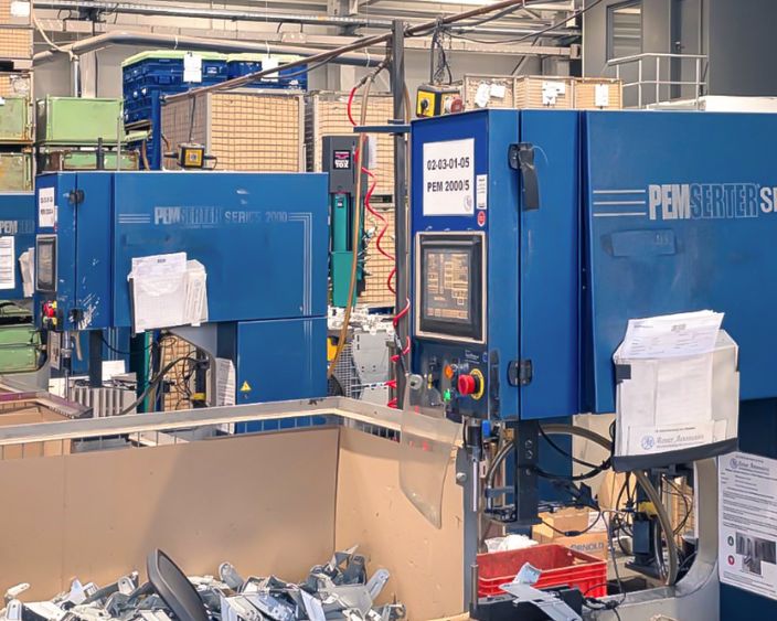 Zwei blaue PEMSERTER® Einpressmaschinen der Serie 2000 in einer Produktionshalle von Alzner Automotive, umgeben von Werkstücken, Kisten und Produktionsmaterialien.