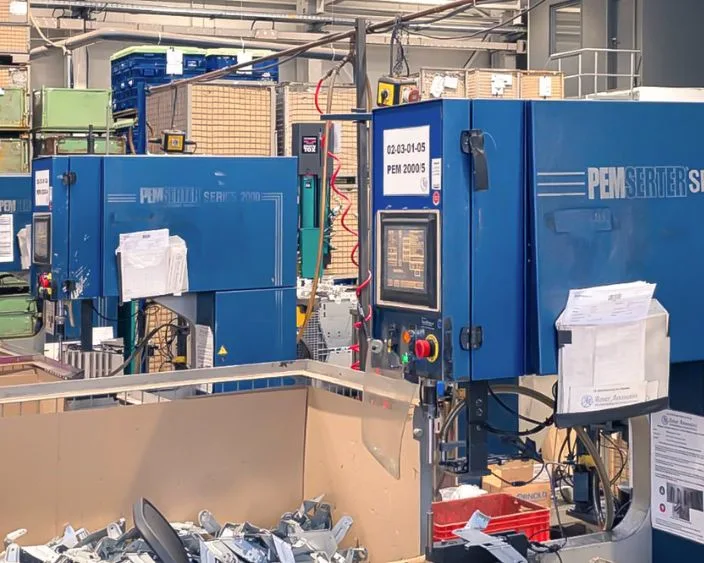 Zwei blaue PEMSERTER® Einpressmaschinen der Serie 2000 in einer Produktionshalle von Alzner Automotive, umgeben von Werkstücken, Kisten und Produktionsmaterialien.
