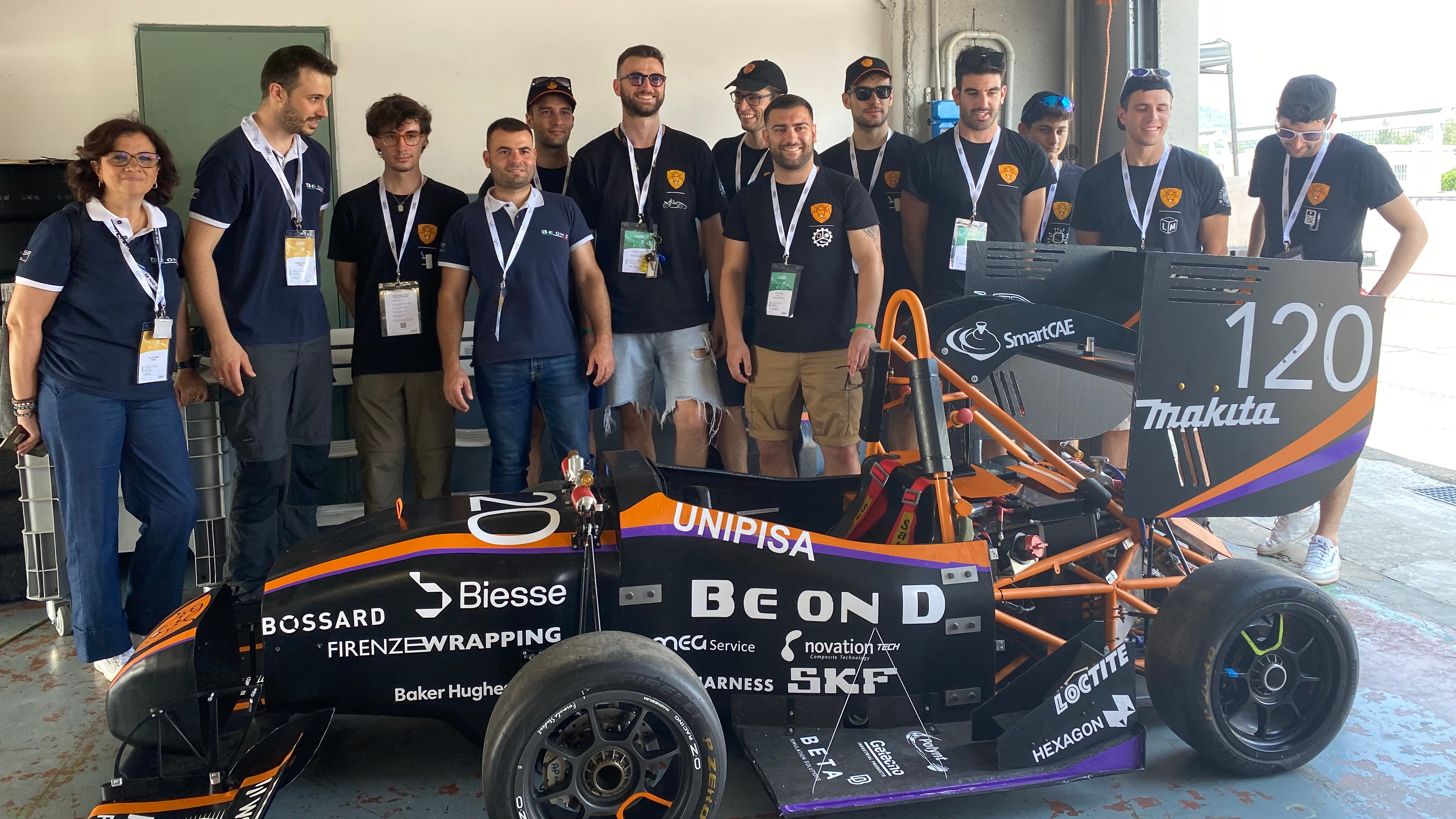 A group of people in matching shirts stand around a black and orange race car with the number 120 and various sponsor logos.