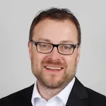 Portrait von Christian Busch mit kurzem Haar, freundlicher Gesichtsausdruck, grauer Hintergrund.