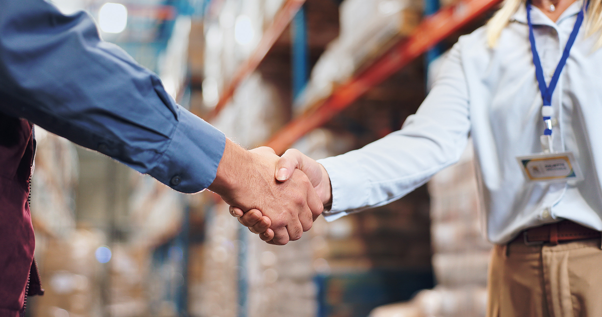 Two people shaking hands in a warehouse, one wearing a blue shirt and the other a white shirt with a name badge.