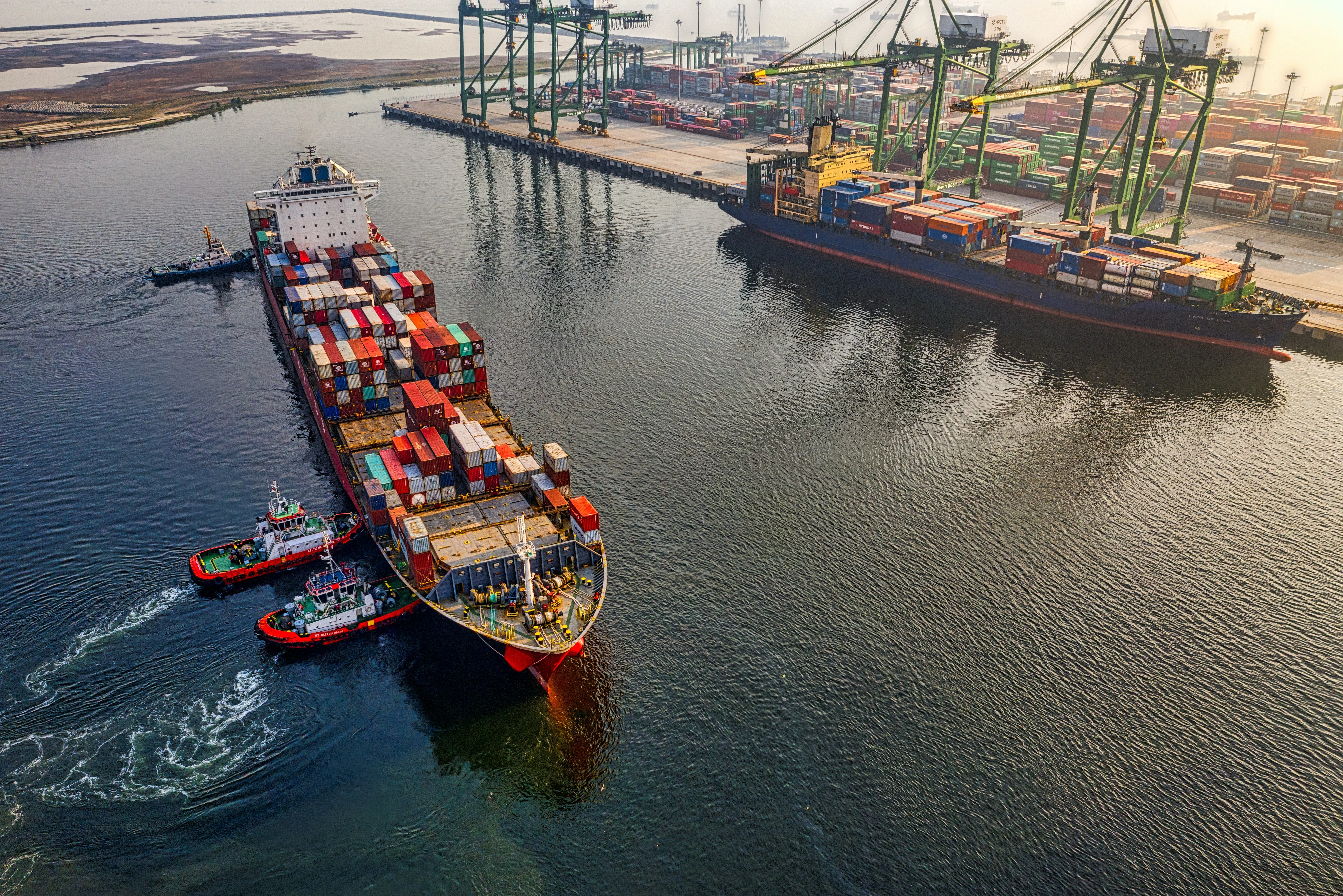 Cargo ship with colorful containers docked at a port, assisted by tugboats, with cranes and stacked containers in the background.