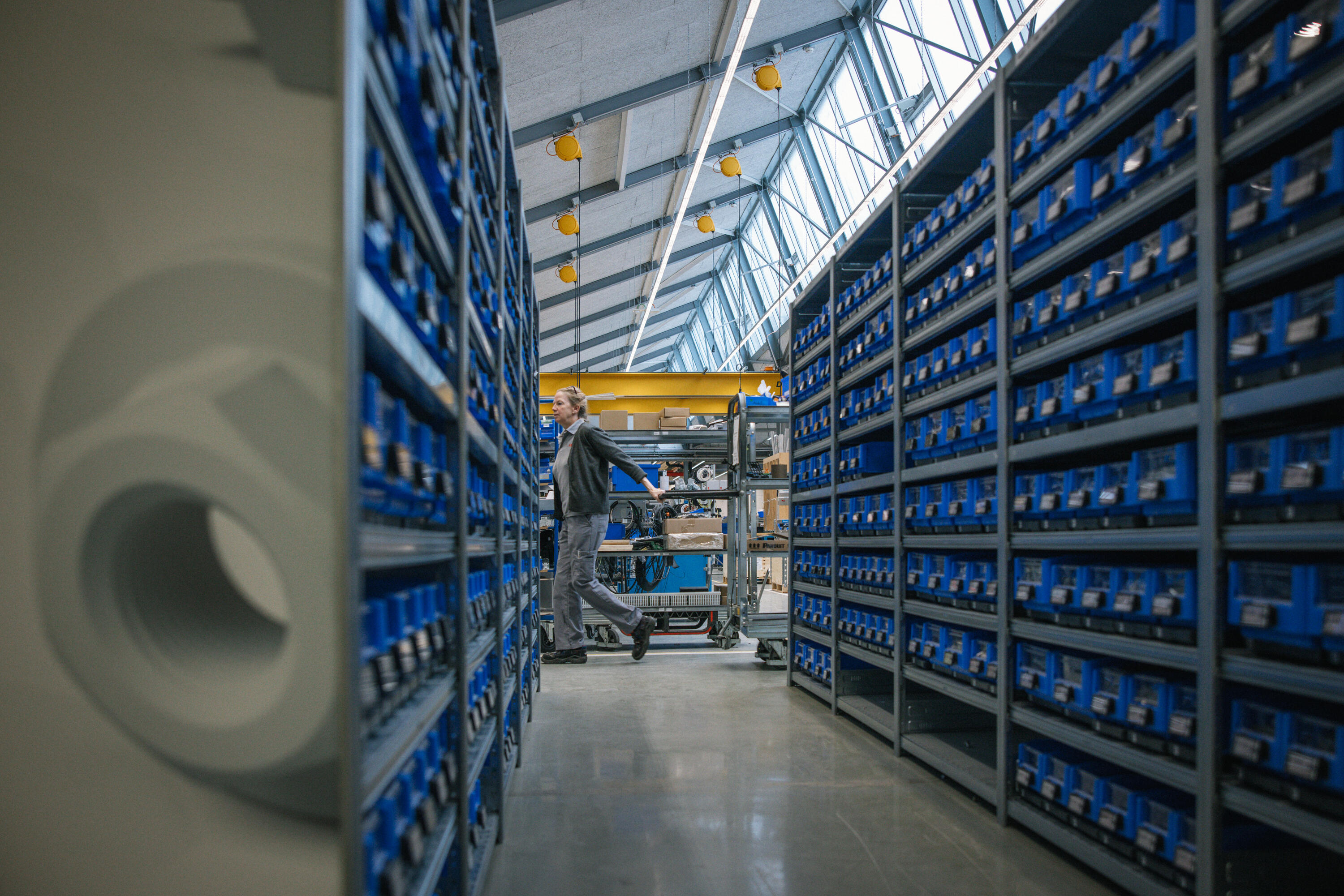 Worker pushing cart through modern warehouse aisle with tall blue storage shelves under glass ceiling with yellow lights.