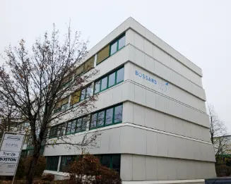Modern white four-story office building with Bossard Aerospace logo and horizontal windows and bare tree in foreground under overcast sky.