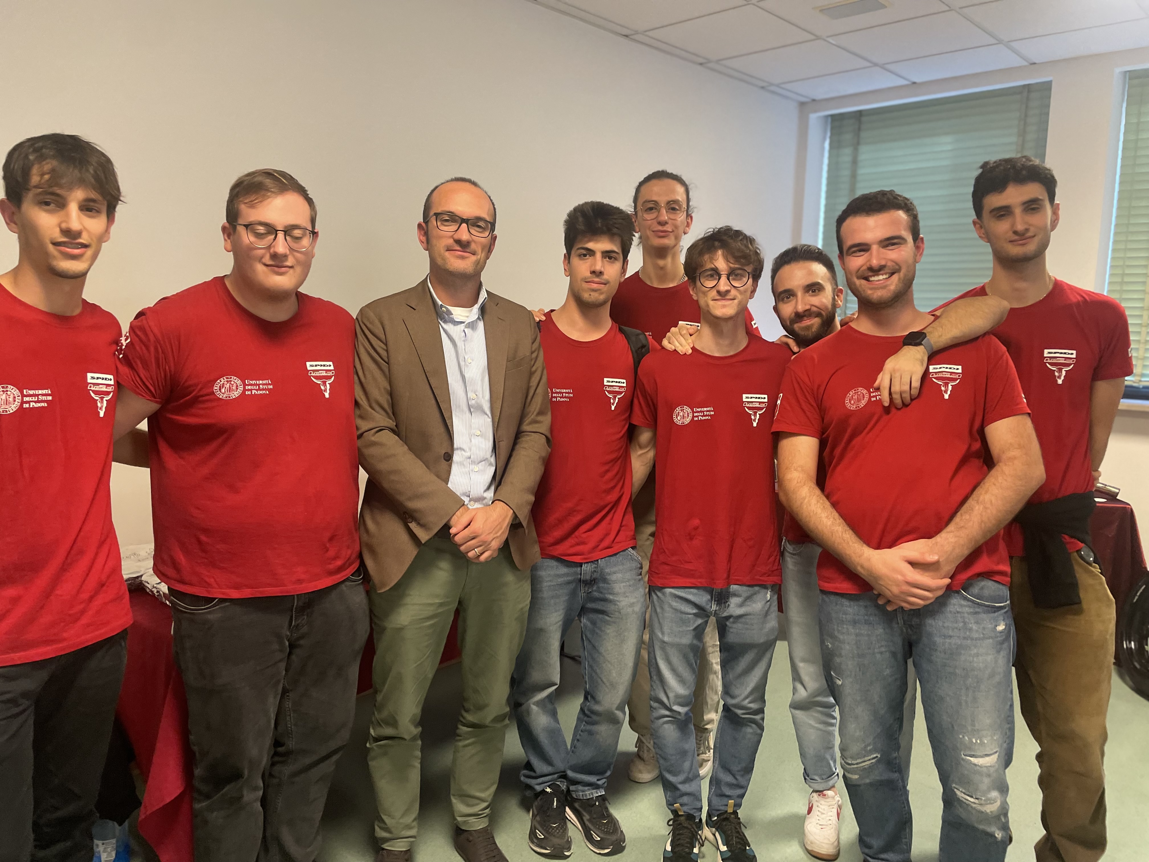 Un gruppo di otto persone, sette con magliette rosse e uno con una giacca marrone, sorride in una stanza con una finestra.