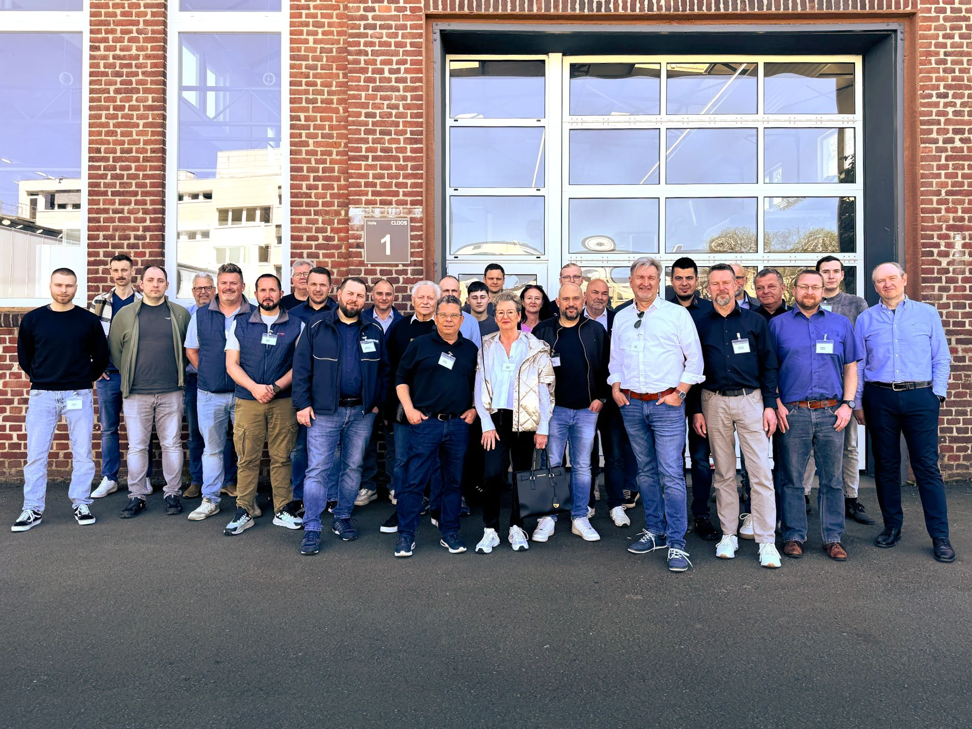 Gruppenfoto mit Teilnehmern der Tagung, mehrere Teilnehmer nebeneinanderstehend vor einer Produktionshalle aus roten Ziegelsteinen mit einer großen Toröffnung aus Glas