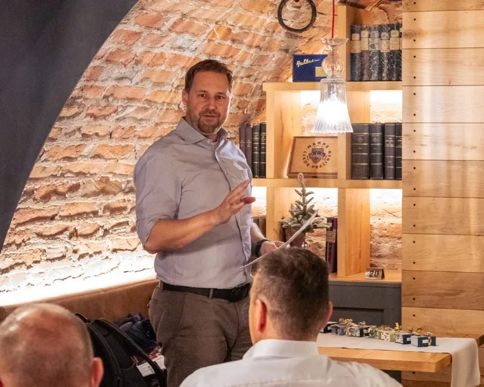 Florian Heuter in einem Gewölbekeller steht vor einer kleinen Gruppe sitzender Personen und spricht. Hinter ihm befinden sich Regale mit Büchern und dekorativen Gegenständen.