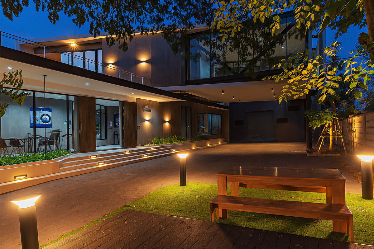Moderne Hausfassade in der Dämmerung mit warmer Beleuchtung, großen Fenstern und einer Holzbank auf einer Rasenfläche, umgeben von Bäumen.
