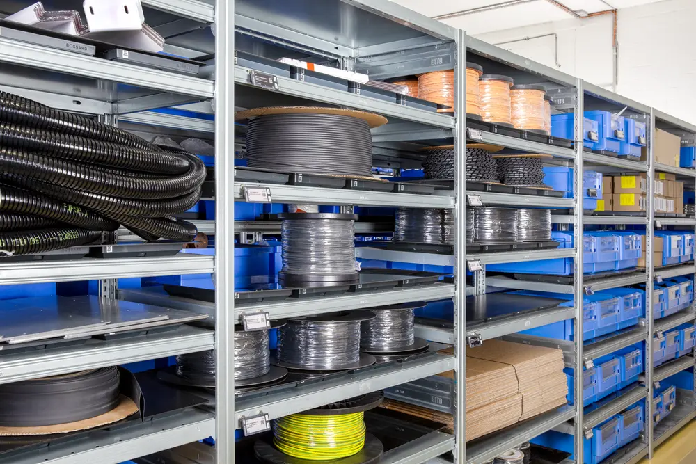 Industrial warehouse shelves with large cable reels, tubing, and blue storage bins organized on metal racks.