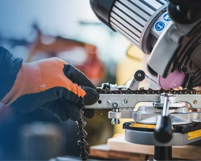 Nahaufnahme einer Kettensägekette beim Schärfen mit einer professionellen Schleifmaschine. Eine behandschuhte Hand fixiert die Kette.