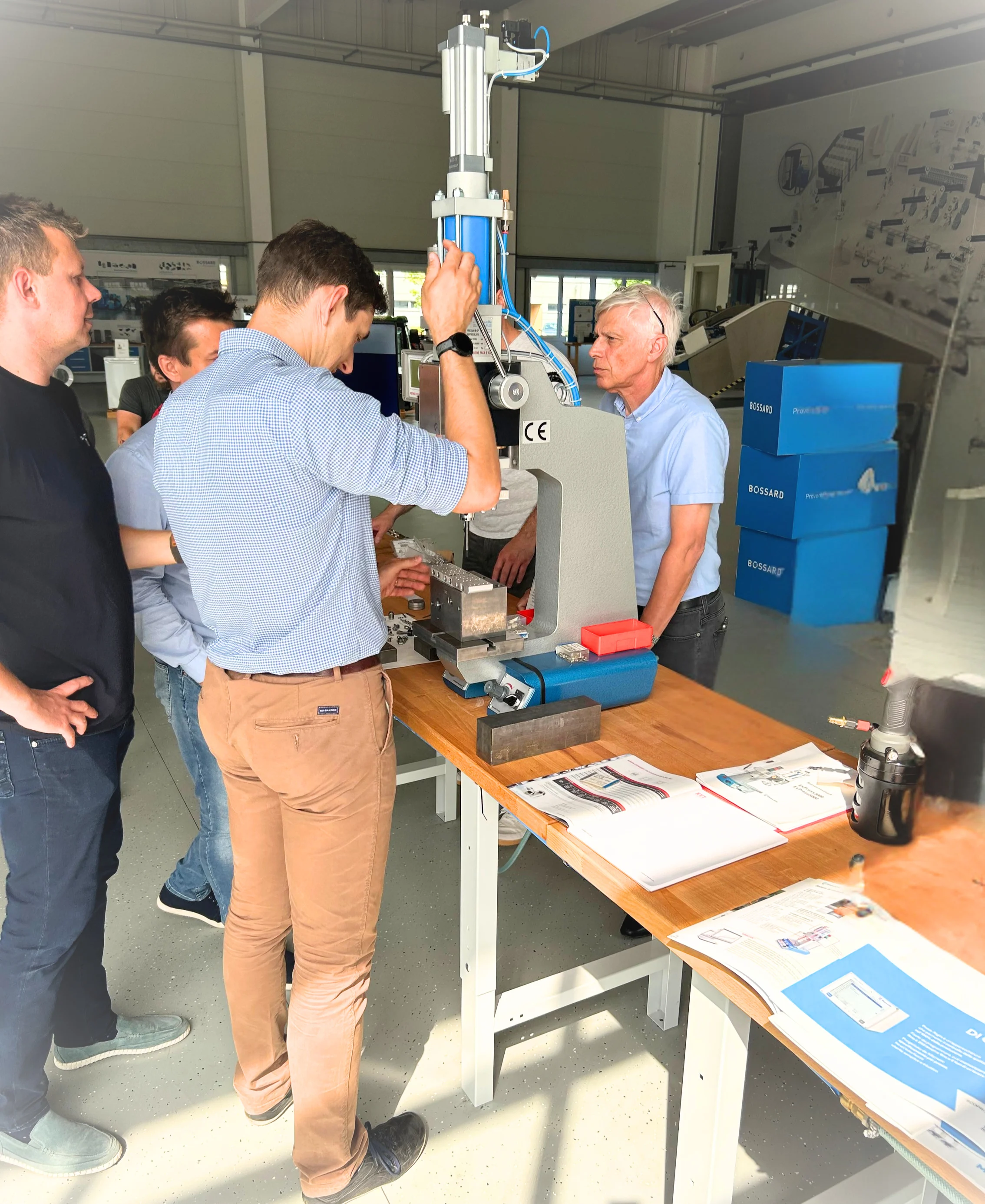 Skupina mužů v Bossard Experience Center v Linci zkoumá bílý a modrý hydraulický lis na stole s technickou dokumentací.