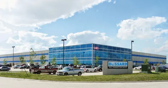 Large industrial building with blue-tinted windows, surrounded by parked cars and a sign reading "Bussard." Cloudy sky above.