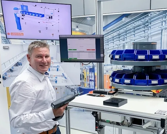 Lächelnder Horst Sälzle in einem Lager mit Barcodescanner vor einem automatisierten Logistiksystem