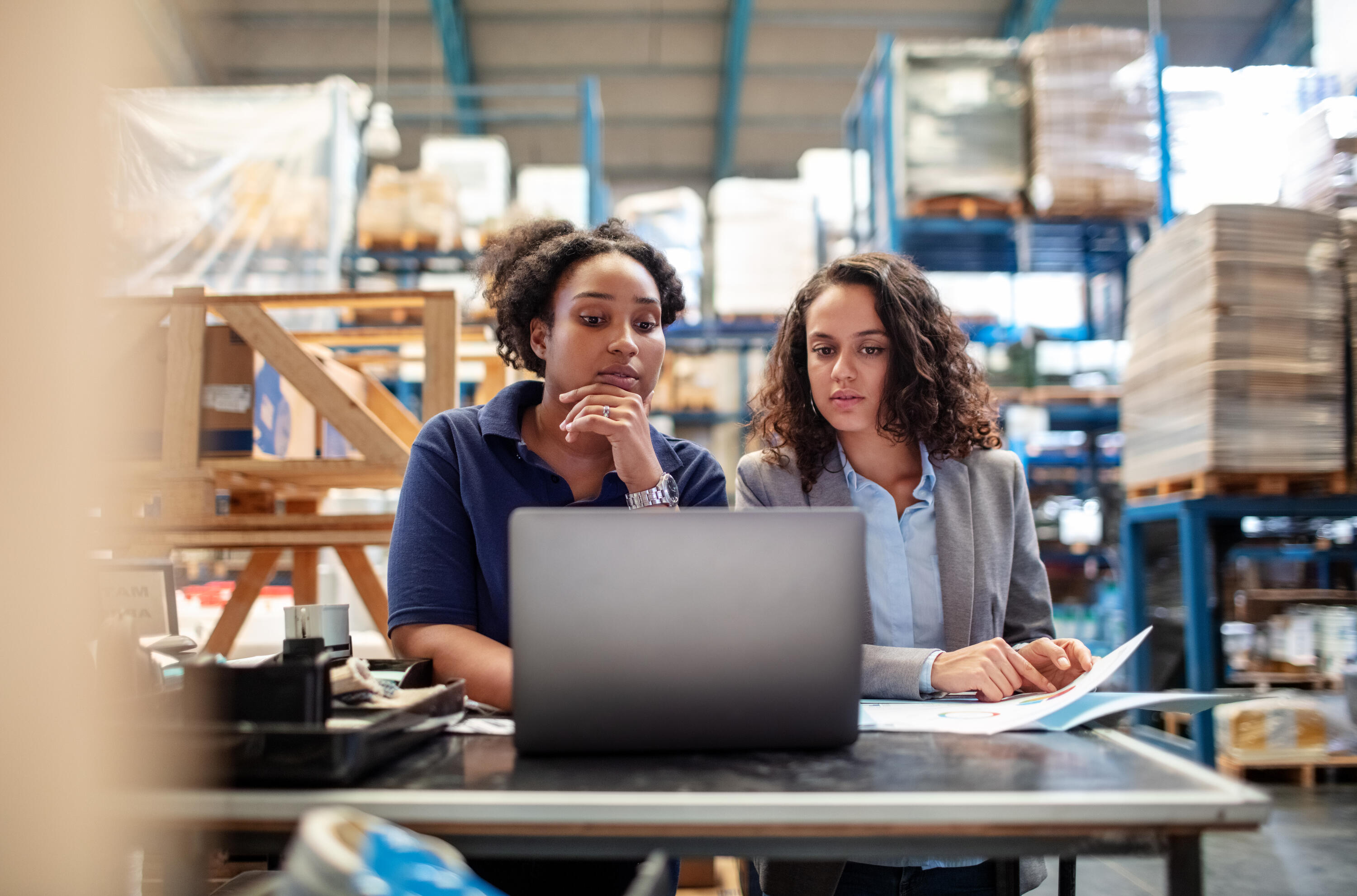 Two women reviewing laptop and documents at desk in warehouse with blue shelving and stacked inventory in background.