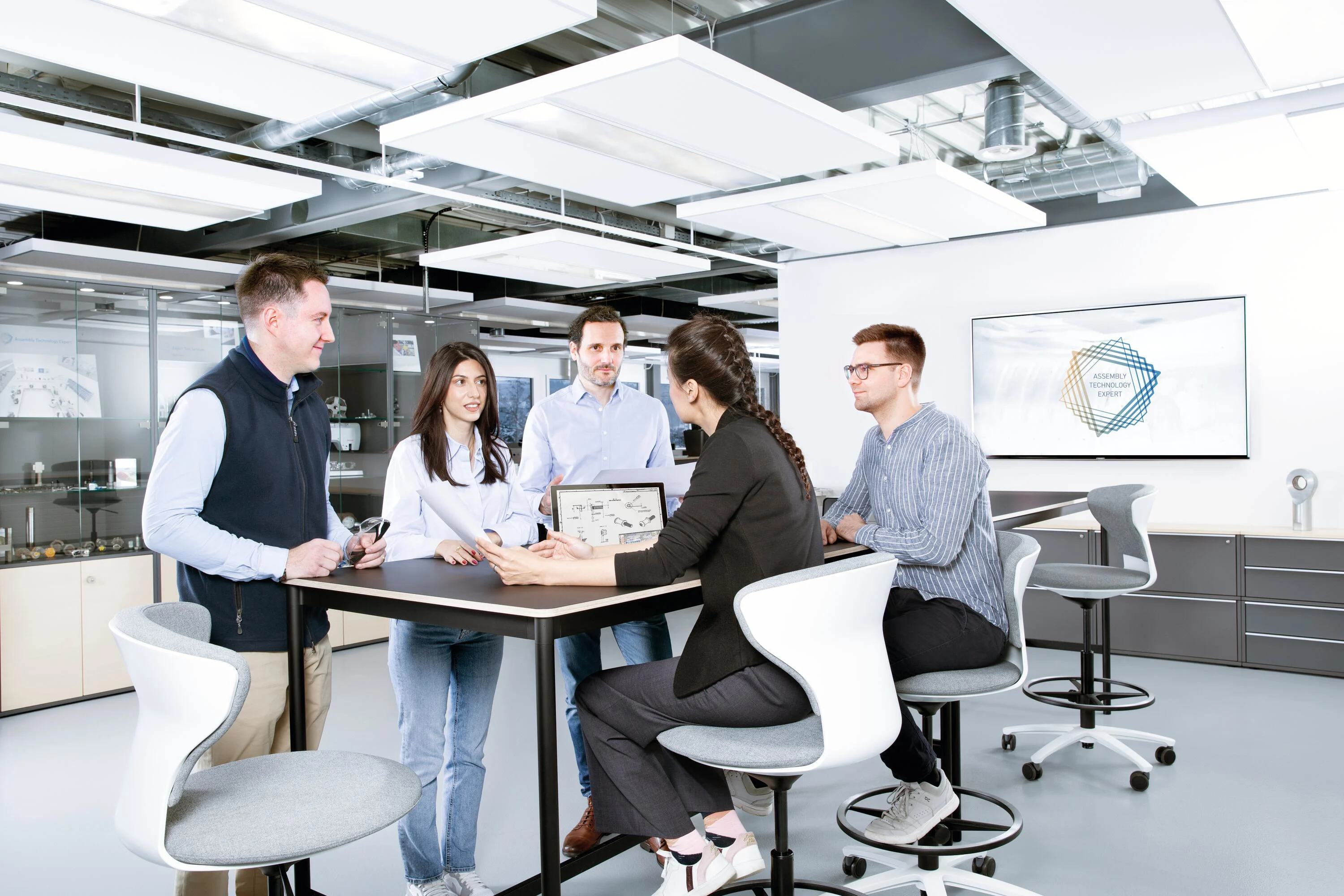 People around a table discussing a technical drawing in an industrial looking office. Key visual for Assembly Technology Expert Service only.
