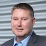 Portrait von Horst Saelzle mit blauem Hemd, leichtes Lächeln, grauer Hintergrund.