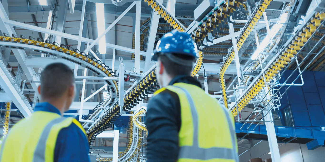 Due uomini indossano gilet ad alta visibilità gialli e osservano la linea di produzione.