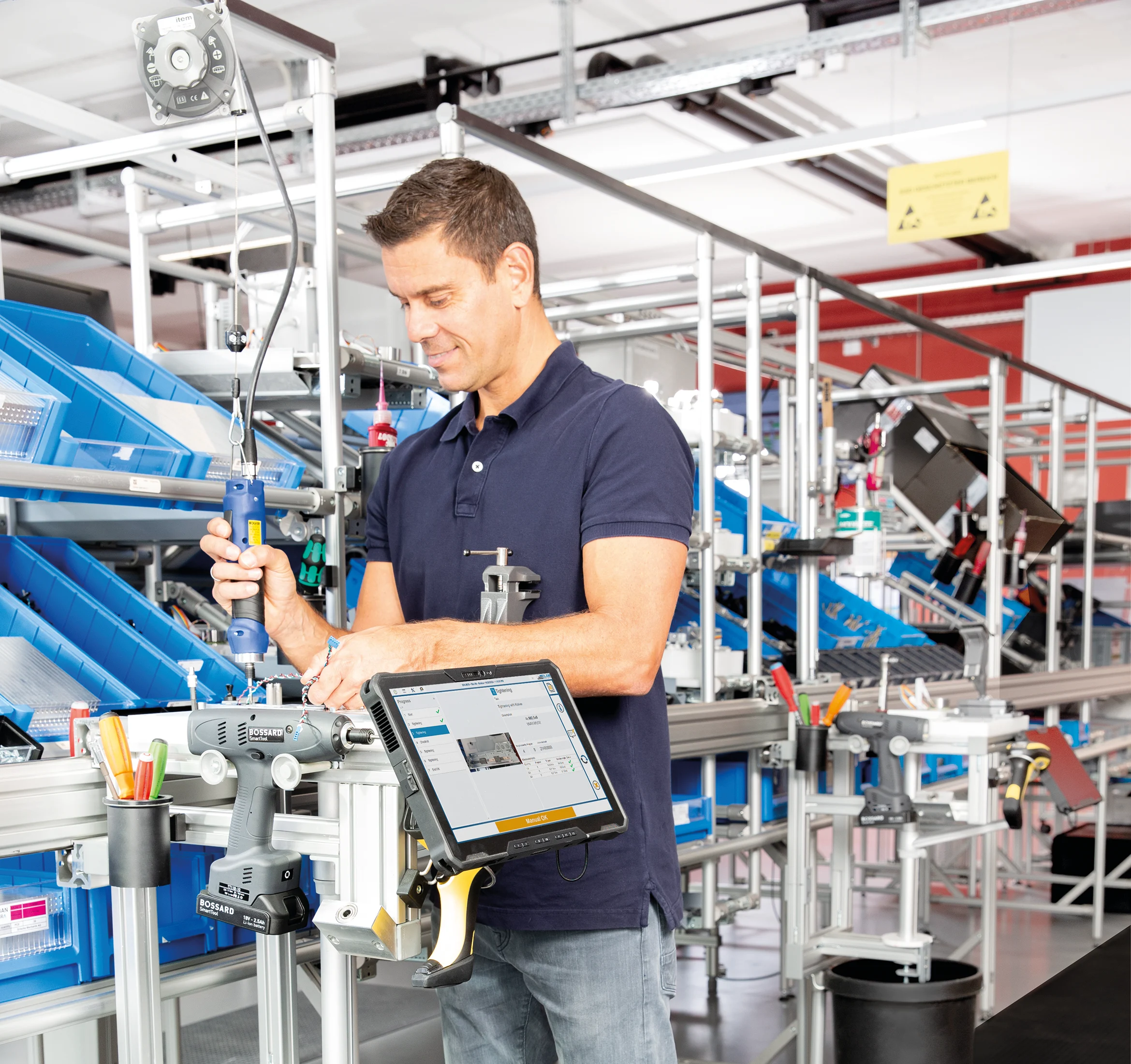 Man in a blue shirt working with tools at an assembly line, surrounded by blue bins and a tablet displaying information.