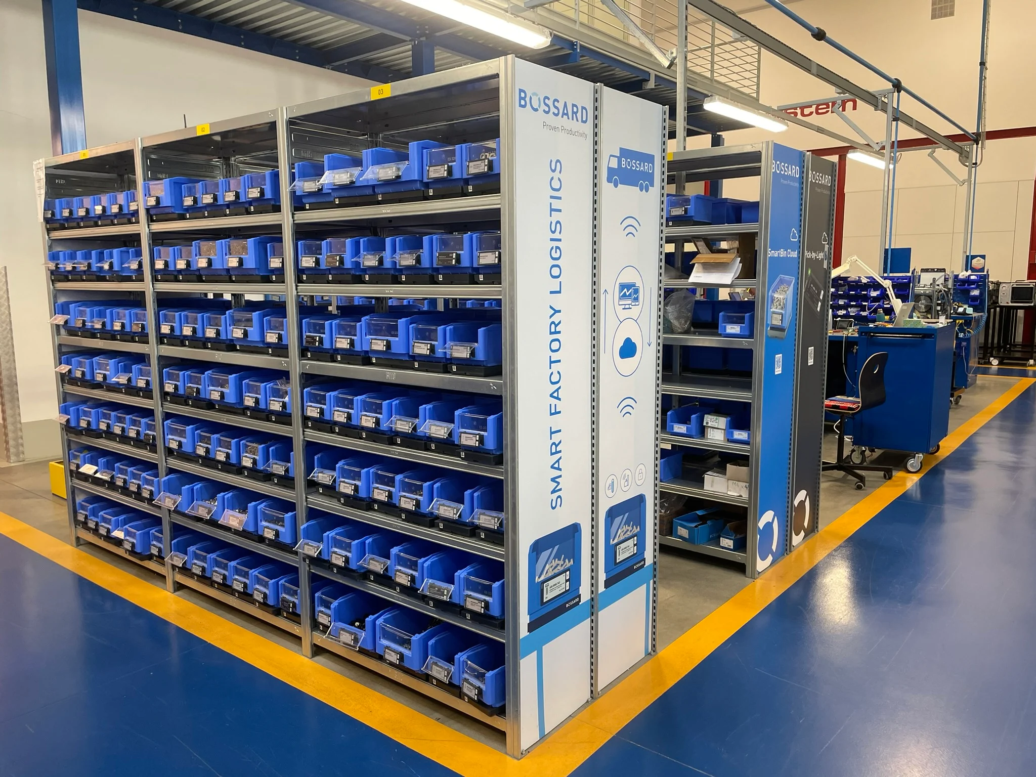 Scaffali di magazzino con contenitori blu etichettati "Smart Factory Logistics" in un ambiente industriale pulito e organizzato.