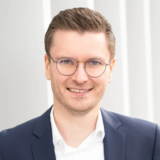 Portrait von Benjamin Hartinger mit Brille, freundliches Lächeln, weißes Hemd mit dunkelblauem Anzug auf hellgrauem Hintergrund.